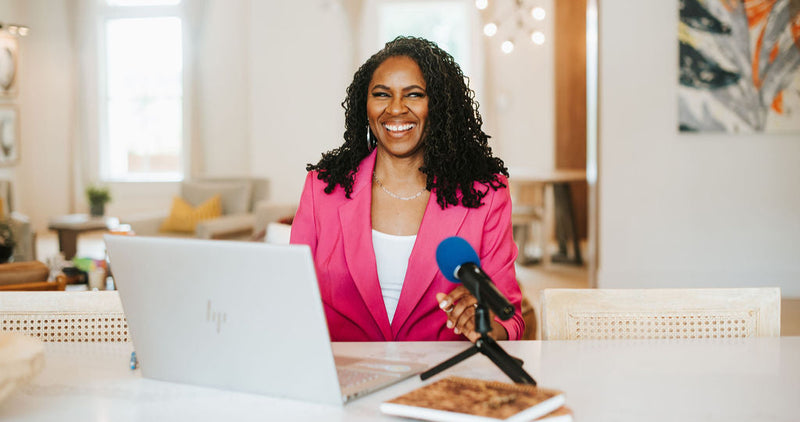 Woman in pink blazer sitting at a desk with a laptop and microphone, smiling.
